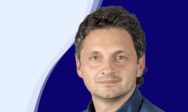 Reinhard Sutterlüty de l'entreprise meusburger sourit à la caméra. Il a les cheveux bouclés et foncés et porte une chemise bleu foncé. L'arrière-plan montre une forme de vague moderne en blanc, bleu clair et bleu foncé, qui s'étend en courbe du bas à gauche au haut à droite de l'image.